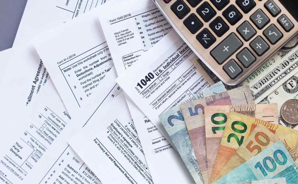 Diversos documentos fiscais, calculadora e notas de diferentes valores sobre uma mesa, representando as preocupações de quem declara imposto de renda e evita cair na malha fina.